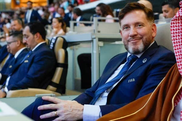 Alejandro Domínguez con traje azul y corbata, con un distintivo de FIFA en la solapa, sentado en una conferencia o evento con una audiencia de fondo. Otros asistentes, también con trajes, están sentados a su alrededor, y una persona con vestimenta tradicional árabe está a su lado.