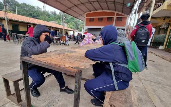 Estudiantes en uniforme azul sentados en bancos de madera bajo una estructura techada en lo que parece ser el patio de una escuela. En el fondo se observan edificios escolares con techos de tejas rojas y otros estudiantes. El ambiente sugiere un recreo o momento de descanso en una institución educativa.